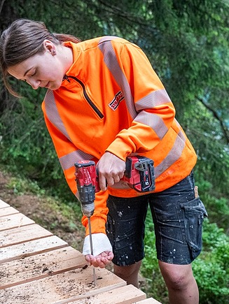 Eine STRABAG Zimmerin Lernender in orangefarbener Sicherheitsjacke arbeitet konzentriert mit einem Akkuschrauber an einer Holzplattform im Wald. Sie befestigt eine Schraube in den Holzdielen, während im Hintergrund dichter Wald und ein See sichtbar sind. 