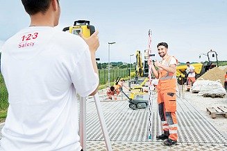 Ein junger STRABAG Lernender mit Helm und Arbeitskleidung steht auf einer Baustelle und arbeitet mit einem Tablet und einem digitalen Stift. Im Hintergrund ist ein eingerüstetes Gebäude sowie Baustellenmaterial zu sehen. 