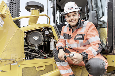 Lehre Baumaschinenmechaniker:in STRABAG Mechaniker Lehrling vor einem Motor einer gelben Baumaschine