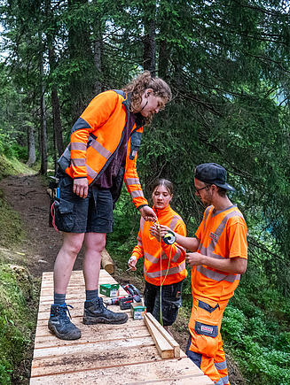 Drei STRABAG Lernende in orangener Sicherheitskleidung arbeiten gemeinsam an einem Holzsteg im Wald. Eine Lernende steht auf dem Steg, während die anderen zwei auf dem Boden Materialien vorbereiten. Die Gruppe nutzt Werkzeuge und Messgeräte, um den Steg präzise auszurichten. Im Hintergrund erstreckt sich dichter Wald, und ein blauer See ist zwischen den Bäumen sichtbar. 
